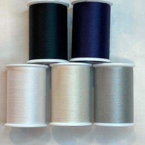 May include: Six spools of thread in various colors: dark green, navy blue, white, cream, and gray. The spools are cylindrical with white plastic bases and tops, and the thread is tightly wound.