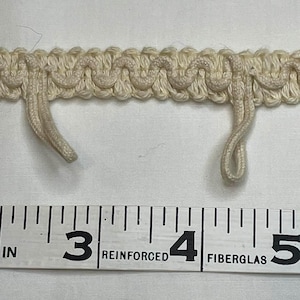 May include: Close-up of a cream-colored decorative trim with a looped design. The trim is measured with a ruler, showing it is approximately 2 inches wide. The trim has two hanging loops.