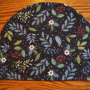May include: A dark, semi-circular fabric item with a festive botanical pattern. The design features green leaves, red berries, and small white flowers on a black background, with white speckles. The item is likely a tea cozy or similar kitchen accessory.