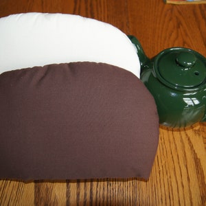May include: Two fabric cushions, one brown and one white, are on a wooden surface. A green teapot is to the right of the cushions.