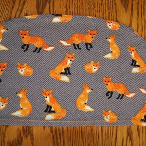 Könnte beinhalten: Ein grauer und orangefarbener Stoff-Tee-Cozy mit einem sich wiederholenden Muster aus Füchsen. Die Füchse sind orange mit schwarzen Augen und Nasen und schauen in verschiedene Richtungen.