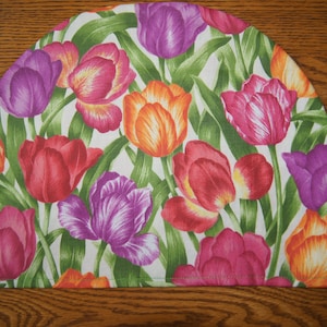 May include: A round, white fabric pot holder with a floral pattern of pink, purple, orange, and yellow tulips. The tulips are on green stems with leaves.