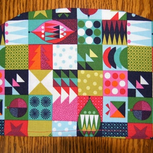 Puede incluir: Un colorido y estampado cubreteiera con forma semicircular. La tela presenta un diseño de patchwork de cuadrados en varios colores, incluyendo rojo, verde, azul y rosa, con formas geométricas y abstractas.