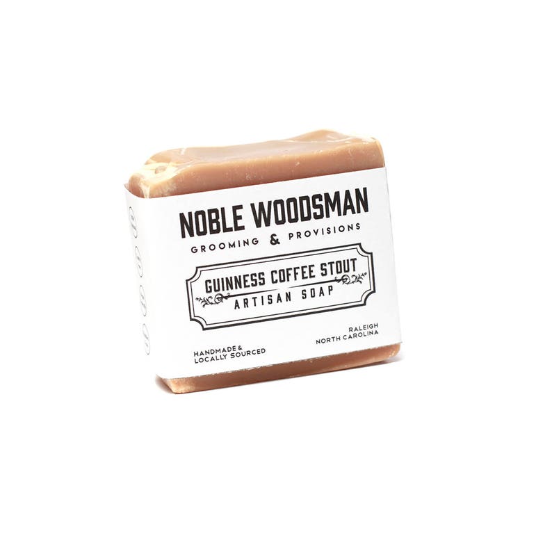 May include: A bar of brown soap with a white label that reads "NOBLE WOODSMAN GROOMING & PROVISIONS GUINNESS COFFEE STOUT ARTISAN SOAP HANDMADE & LOCALLY SOURCED RALEIGH NORTH CAROLINA"