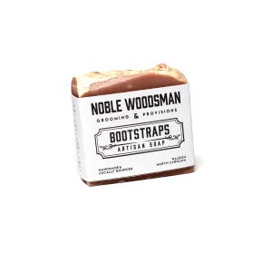 May include: A bar of brown soap with white lettering that reads "NOBLE WOODSMAN GROOMING & PROVISIONS BOOTSTRAPS ARTISAN SOAP HANDMADE & LOCALLY SOURCED RALEIGH NORTH CAROLINA"