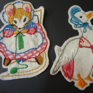 May include: Two embroidered patches: a mouse knitting and a goose. The mouse wears a blue cape and a white dress with red trim. The goose has a blue bonnet and a red bow. Both are on a white felt background.