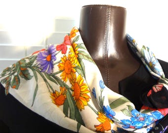 May include: A floral scarf with a cream background and a colorful sunflower and floral pattern. The scarf is draped around a mannequin's neck, showcasing the vibrant design. The scarf features orange, yellow, blue, purple, and red flowers.