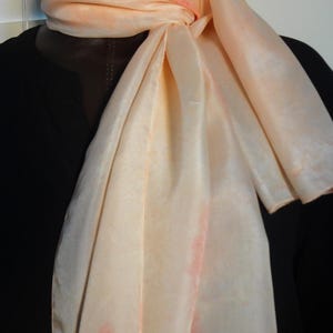 May include: A light peach-colored silk scarf with a subtle ombre effect. The scarf is tied around a mannequin's neck and has the word "Sophya" embroidered in green on the bottom right corner.
