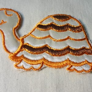May include: A white and brown embroidered turtle applique patch. The turtle has a brown shell with white stripes and a white head and legs.