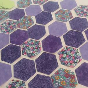 May include: A collection of purple and floral fabric hexagons, arranged in a honeycomb pattern. The hexagons are various shades of purple, with some featuring floral patterns.
