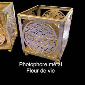 Könnte beinhalten: Zwei goldene Kerzenhalter aus Metall mit geometrischem Design. Die Kerzenhalter sind quadratisch und haben einen runden Ausschnitt in der Mitte. Der Text "Photophore métal Fleur de vie" ist unter den Kerzenhaltern gedruckt.