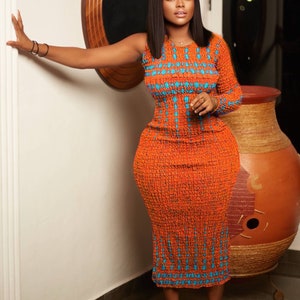 May include: A woman wearing a long, orange dress with a blue and white geometric pattern. The dress has one sleeve and is fitted to the body. She is standing in a room with a white wall and a wooden door.