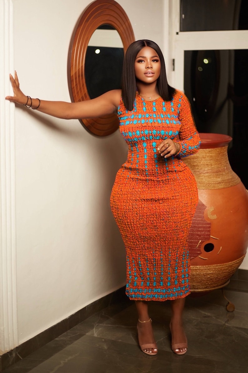 May include: A woman wearing a long, orange dress with a blue and white geometric pattern. The dress has one sleeve and is fitted to the body. She is standing in a room with a white wall and a wooden door.