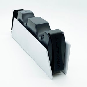 Puede incluir: Un soporte blanco y negro para consola de juegos con un soporte negro para mandos. El soporte tiene el texto "VIMMOUNT" impreso en el lateral. El soporte está diseñado para sujetar varios mandos.