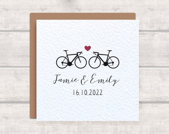 May include: A white card with two black bicycles facing each other with a small red heart above them. The text "Jamie & Emily" and "16.10.2022" is below the bicycles.