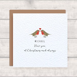Könnte beinhalten: Quadratische Grußkarte mit braunem Rand. Die Karte zeigt zwei Vögel, die sich mit einem roten Herzen über ihnen gegenüberstehen. Der Text lautet: "MICHAEL Ich liebe dich, zu Weihnachten und immer."