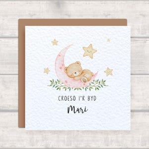 Könnte beinhalten: Eine quadratische Grußkarte mit einer Aquarellillustration eines braunen Teddybären, der auf einem rosa Halbmond schläft, umgeben von grünen Blättern und gelben Sternen. Der Text lautet CROESO I'R BYD und der Name Mari in Schwarz.