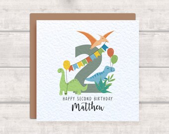 May include: A birthday card with a green, blue, and orange dinosaur design. The number 2 is in the center of the card with a pterodactyl flying above it. The card says "Happy Second Birthday Matthew".
