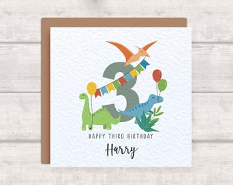 May include: A birthday card with a green, blue, and orange dinosaur theme. The card features a large number 3 with a dinosaur flying above it. The text "Happy Third Birthday Harry" is printed on the card.