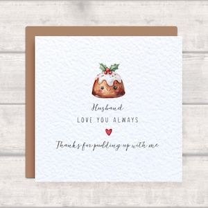 Könnte beinhalten: Quadratische Karte mit braunem Rand. Sie zeigt eine Aquarellillustration eines lächelnden Weihnachtspuddings mit Stechpalmen und Beeren. Der Text lautet: "Husband, Love You Always, Thanks for pudding up with me."
