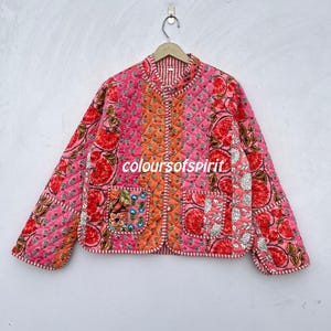 May include: Pink quilted jacket with floral embroidery and orange accents. The jacket features a round collar, two pockets, and the text "coloursofspirit". The jacket is hanging on a wooden hanger.