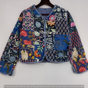 May include: A blue quilted jacket with a floral patchwork design. The jacket has a V-neck and long sleeves. The fabric is quilted and has a textured surface. The jacket is trimmed with white piping. The text "COLOURSOF SPIRIT" is visible on the front of the jacket.