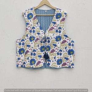 Peut inclure: Un gilet matelassé blanc avec un motif floral bleu et une doublure rayée bleu et blanc. Le gilet a une fermeture à nouer avec un pompon. Le texte "COLOURSOFSPIRIT" est imprimé sur le devant du gilet.