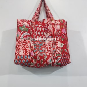 以下が含まれることがあります： 白、緑、ピンクの花柄が施された赤いキルトトートバッグ。バッグには2つのハンドルがあり、前面には「COLOURSOFSPIRIT」の文字がプリントされています。バッグは布製で、パッチワークのデザインです。