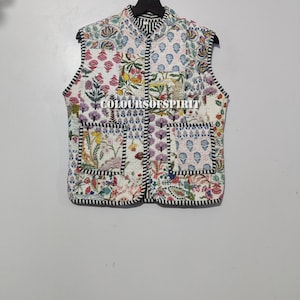 May include: A white quilted vest with a black and white striped collar and trim. The vest has a patchwork design with floral patterns in shades of pink, purple, blue, and green. The vest has two pockets on the front.