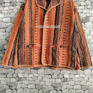 May include: A long-sleeved jacket featuring a quilted texture with vertical stripes in shades of orange, brown, and black. The garment has a wide collar, two front pockets, and white running stitch details throughout. The text coloursofspirit is visible.