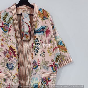 K&ouml;nnte beinhalten: Eine rosa Kimono-Robe mit floralem Druck, einem gestreiften Saum und einem passenden rosa G&uuml;rtel mit floralem Druck. Die Robe hat lange &Auml;rmel und einen V-Ausschnitt.