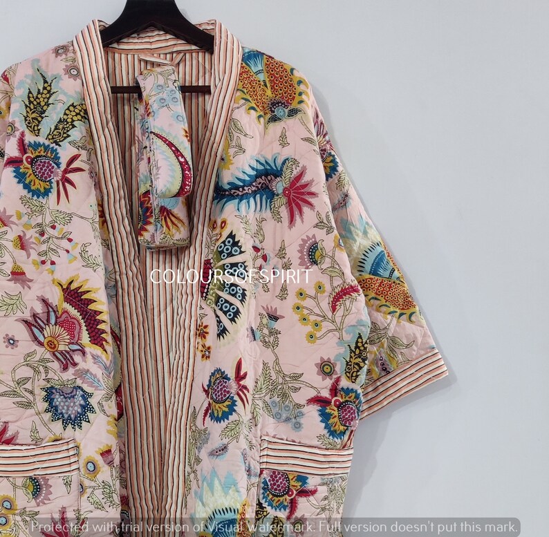 K&ouml;nnte beinhalten: Eine rosa Kimono-Robe mit floralem Druck, einem gestreiften Saum und einem passenden rosa G&uuml;rtel mit floralem Druck. Die Robe hat lange &Auml;rmel und einen V-Ausschnitt.