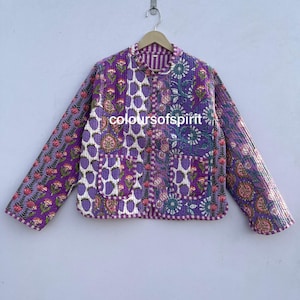 May include: A quilted jacket with a patchwork design in shades of purple, pink, and white. The jacket features floral and geometric patterns, a round collar, and two pockets. The text "coloursofspirit" is visible on the jacket.