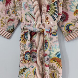 K&ouml;nnte beinhalten: Eine rosa gesteppte Robe mit Blumenmuster und gestreiftem Saum. Die Robe ist mit einem passenden G&uuml;rtel in der Taille gebunden. Der Text "COLOURS OF SPIRIT" ist auf die Robe gedruckt.