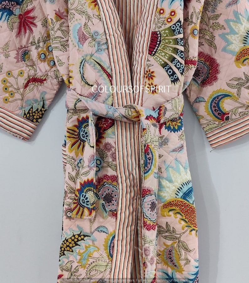 K&ouml;nnte beinhalten: Eine rosa gesteppte Robe mit Blumenmuster und gestreiftem Saum. Die Robe ist mit einem passenden G&uuml;rtel in der Taille gebunden. Der Text "COLOURS OF SPIRIT" ist auf die Robe gedruckt.