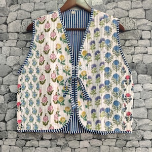 May include: A sleeveless quilted waistcoat featuring a white base with vertical floral patterns in pink, yellow, and blue. The garment has a V-neckline, a front tie closure, and blue and white striped trim. The interior lining is blue and white striped.