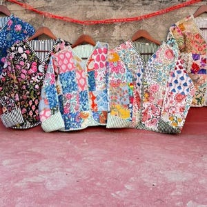 May include: Five quilted jackets with V-necklines and long sleeves, each featuring distinct patchwork patterns in various floral and geometric prints. The jackets are displayed on wooden hangers against a textured wall.