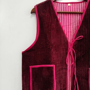 Può includere: Un gilet senza maniche in velluto bordeaux scuro con cuciture trapuntate verticali e bordini rosa acceso. La parte anteriore presenta una chiusura a laccio e due tasche applicate. La fodera interna è in tessuto a righe verticali bianche e rosa.