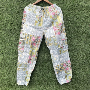 May include: Light blue pants featuring a multi-colored floral and paisley block print pattern. The pants have an elasticized waistband with a drawstring tie, side cargo pockets, and a relaxed fit, hanging from a wooden clothes hanger.