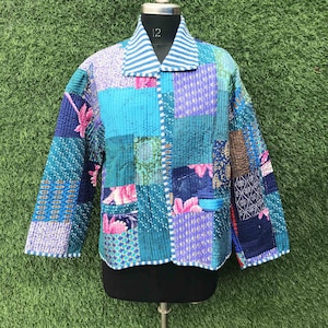 May include: A quilted jacket featuring a patchwork design of various blue, teal, and purple fabrics with floral and geometric patterns. The jacket has long sleeves, a collared neckline with white and blue stripes, and a front button closure.