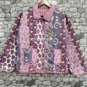 May include: A quilted jacket featuring a patchwork design of purple, lavender, and white floral and strawberry patterns. The garment has a collared neckline, long sleeves, a button-front closure, and two front patch pockets. The text "coloursofspirit" is visible.