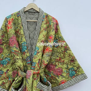 Könnte beinhalten: Eine gesteppte grüne Robe mit einem Blumenmuster in Rot, Rosa und Türkis. Die Robe hat schwarz-weiß gestreifte Verzierungen an Kragen, Manschetten und Taschen. Der Text "coloursofspirit" ist sichtbar.