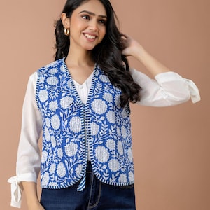 Può includere: Un gilet trapuntato senza maniche con un vivace motivo floreale blu e bianco stampato a blocchi. Il gilet presenta uno scollo a V, una chiusura frontale a laccio ed è rifinito con un sottile bordo a righe blu e bianche lungo le aperture.