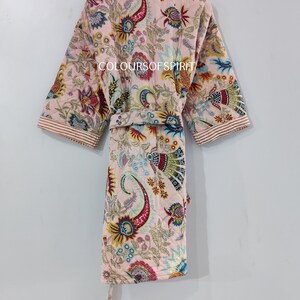 K&ouml;nnte beinhalten: Eine rosa gesteppte Robe mit einem Blumendruck in verschiedenen Farben, darunter Rot, Blau, Gelb und Gr&uuml;n. Die Robe hat einen Selbstg&uuml;rtel und einen Kragen. Der Text "COLOURSOF SPIRIT" ist auf die Robe gedruckt.
