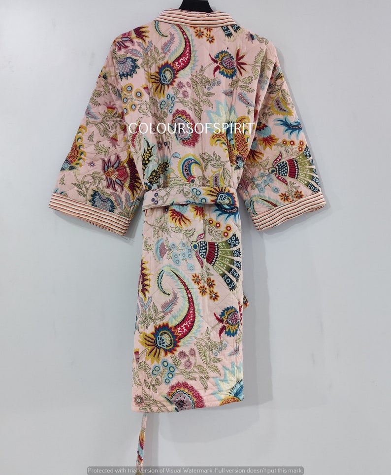 K&ouml;nnte beinhalten: Eine rosa gesteppte Robe mit einem Blumendruck in verschiedenen Farben, darunter Rot, Blau, Gelb und Gr&uuml;n. Die Robe hat einen Selbstg&uuml;rtel und einen Kragen. Der Text "COLOURSOF SPIRIT" ist auf die Robe gedruckt.
