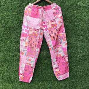 Könnte beinhalten: Rosa Patchwork-Cargohose mit einer Vielzahl von Blumen-, Paisley- und geometrischen Mustern in Magenta-, Hellrosa- und Weißtönen. Die Hose verfügt über einen elastischen Bund mit Kordelzug sowie seitliche Cargotaschen an den Beinen.