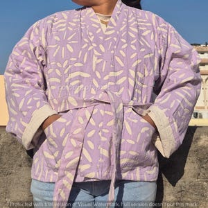 May include: A light purple kimono-style jacket with a white leaf pattern. The jacket has a matching belt, pockets, and cream-colored cuffs. The jacket is made of a soft, quilted fabric.
