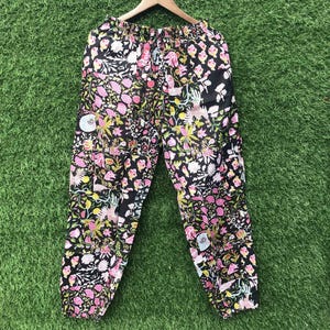 May include: Black trousers featuring an all-over floral print in shades of pink, yellow, and white. The trousers have an elasticated waistband with a drawstring closure and elasticated cuffs at the ankles, displayed on a wooden clothes hanger.