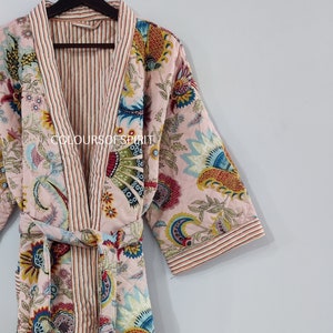 K&ouml;nnte beinhalten: Eine rosa gesteppte Robe mit Blumenmuster und rot-wei&szlig; gestreiftem Besatz. Die Robe hat einen Bindeg&uuml;rtel und lange &Auml;rmel. Der Text "COLOURS OF SPIRIT" ist auf der Robe sichtbar.