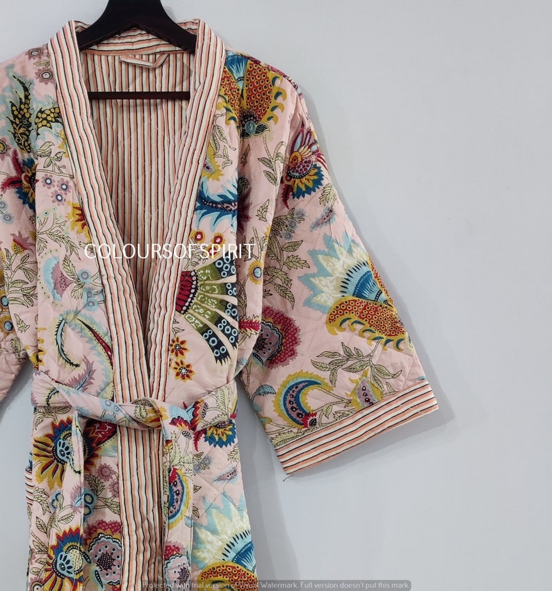 K&ouml;nnte beinhalten: Eine rosa gesteppte Robe mit Blumenmuster und rot-wei&szlig; gestreiftem Besatz. Die Robe hat einen Bindeg&uuml;rtel und lange &Auml;rmel. Der Text "COLOURS OF SPIRIT" ist auf der Robe sichtbar.